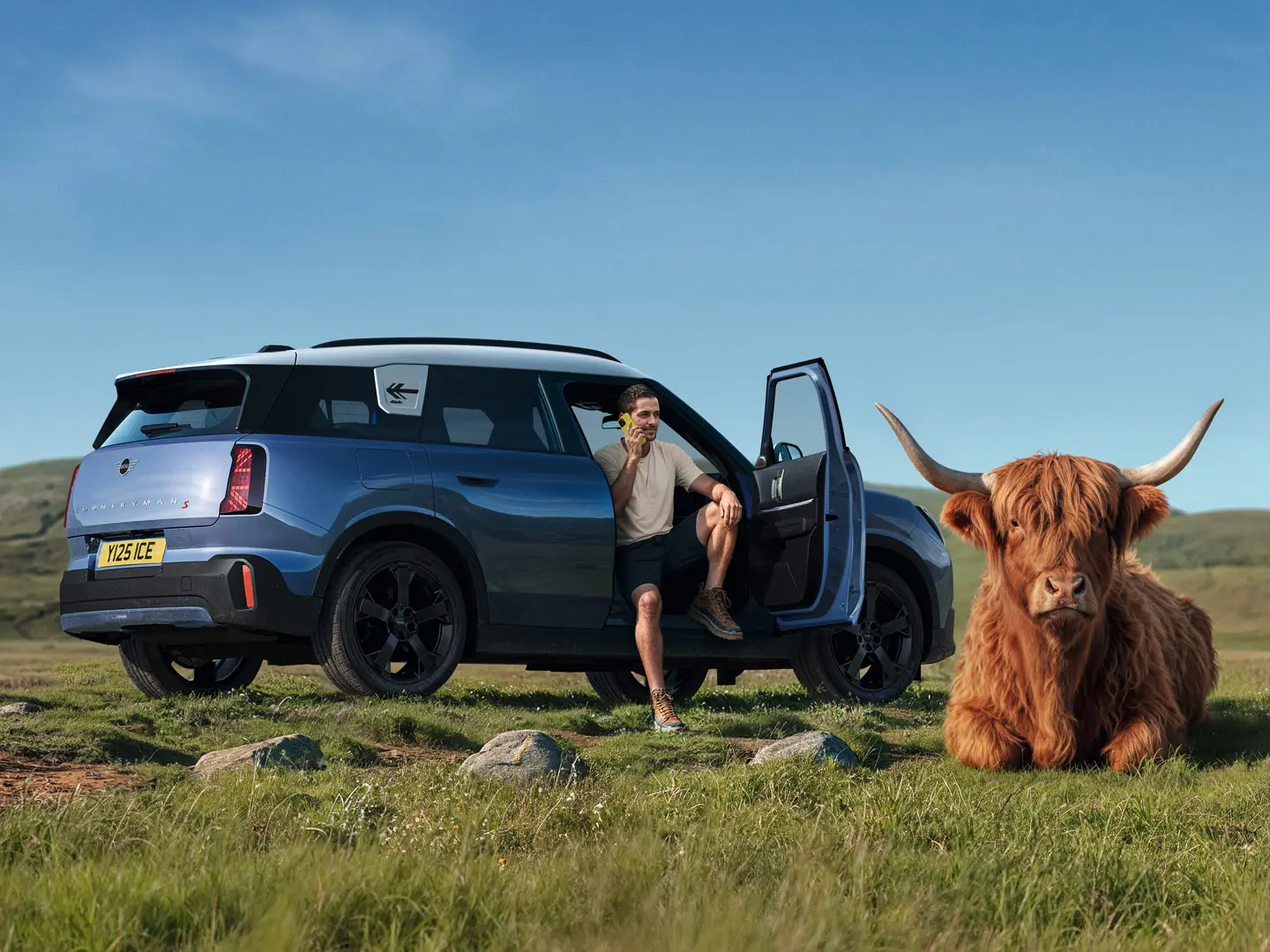 Man outside his MINI Countryman S talking to MINI Roadside Assistance via the MINI App with a Highland Bull lying nearby.