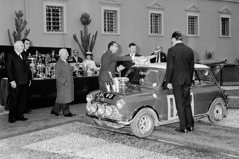 The BMC Works Mini Cooper S wins the 1965 Monte Carlo Rally. Trophies are awarded in front of a historical building.